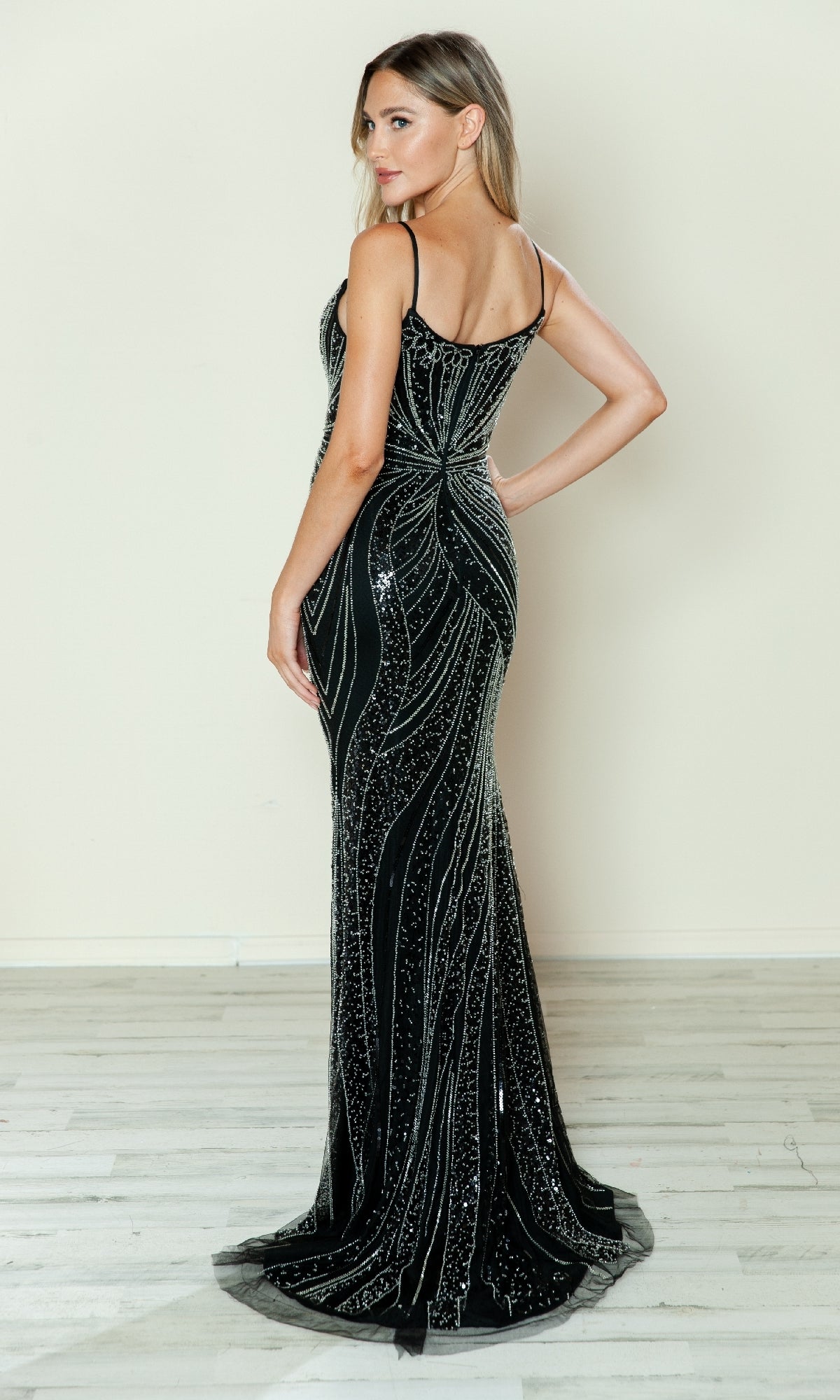 A woman stands sideways on a light wood floor against a beige wall, wearing the Poly USA 9510 Beaded Spaghetti-Strap Prom Gown—a sleeveless, floor-length black dress adorned with silver sequins.